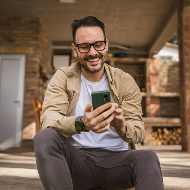 um homem adulto na casa da frente no dia usar chamada de vídeo do telefone inteligente ou sms - mid adult men smart casual white happiness - fotografias e filmes do acervo