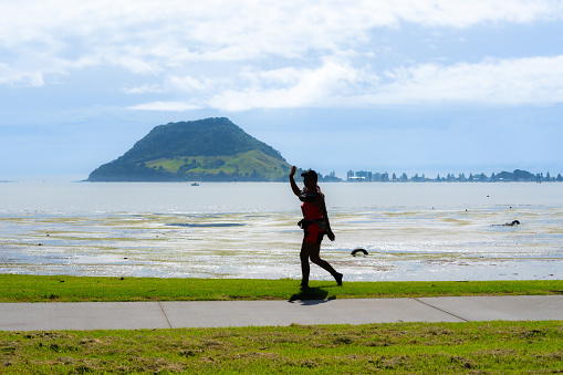 Tauranga New Zealand - April 2 2023; Woman in silhouette walks briskly along waterfront path with landmark Mount Maunganui in background.