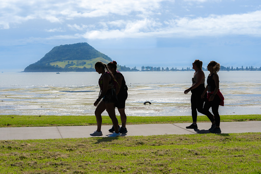 Tauranga New Zealand - April 2 2023; Four women in twos and in silhouette walking along harbour side walkway with Mount Maunganui in background