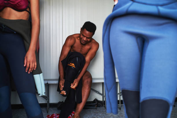 Front view of a sitting young surfer putting on his wetsuit in the dressing room.