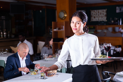 Portrait of polite asian waitress with serving tray warmly welcoming in cozy modern restaurant