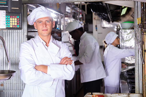 Confident chef of restaurant posing in kitchen on background with working employees