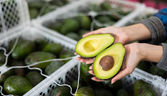 Farmer woman holding cut ripe fleshy hass avocado in hands against stack of boxes with sorted avocados, cropped shot