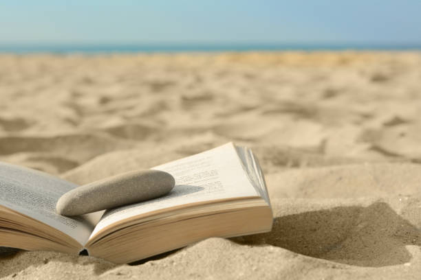 istock Open book with stone on sandy beach near sea, closeup. Space for text 1479858341