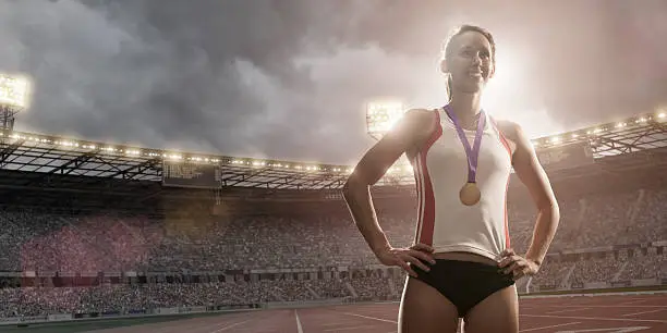 Happy female athlete standing in floodlit athletics stadium wearing gold medal