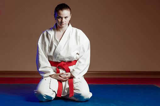 Female portrait of young woman wear white karategi who practising karate indoors. Girl exercising sports training in the gym on tatami mat. Lifestyle with people and martial arts, color neon red and blue lighting. Person with belt sitting on floor