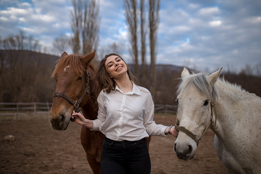 Beautiful white woman holding reins of her brown and white horse and leading them. She s between two horses. In the background blue sky with white clouds.