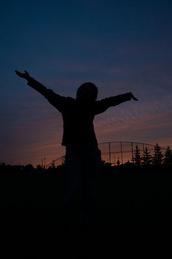 The silhouette of a woman with her arms in the air at a park during dusk with colorful clouds above