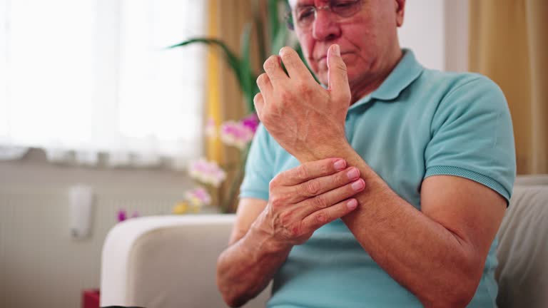 Senior  man rubbing his hands in discomfort, suffering from arthritis in his hand while sitting on sofa at home. Elderly and health issues concept