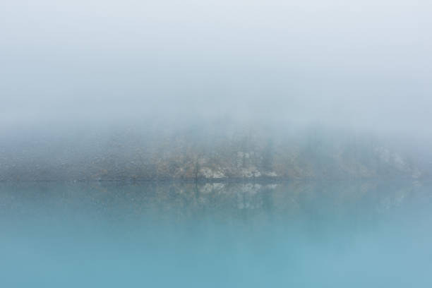 istock Simple meditative landscape with turquoise lake in thick fog. Tranquil view through clearance in dense fog to steep slope in fading autumn colors and mirror mountain lake. Reflective azure alpine lake 1479880068