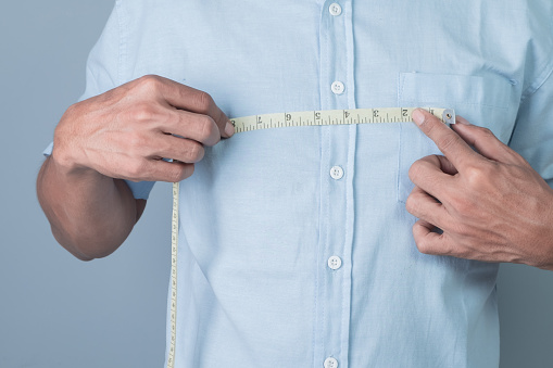A man measures the width of his chest with a tape measure on a gray background.