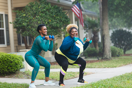 Two African-American women exercising together on the sidewalk in front of a house. They are doing squats with hand weights. The plus size woman with dyed hair is in her 30s. Her friend or personal trainer is a mature woman in her 40s.
