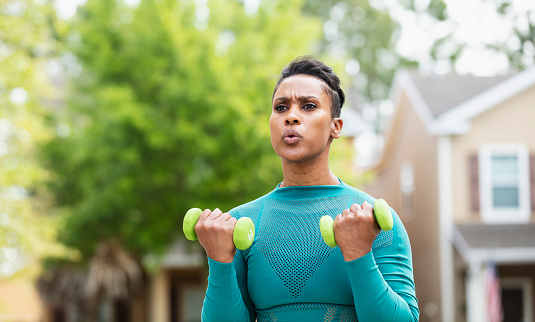 Headshot of a mature African-American woman in her 40s exercising outdoors, houses out of focus in the background. She is looking away with a serious expression, concentrating, doing bicep curls.