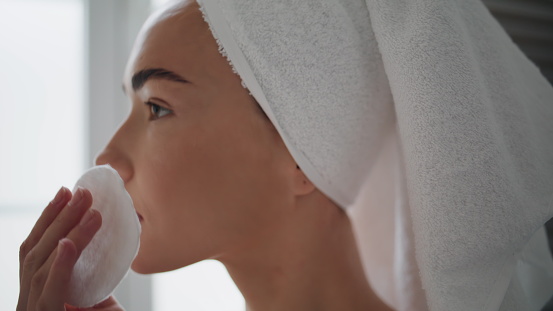 Gorgeous woman cleaning face in bathroom closeup. Serious girl rubbing skin with cotton disposable pad doing morning routine. Satisfied lady with towel on head removing impurities after shower at home