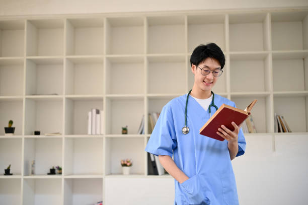 istock Smart young Asian male medical student reading a book while standing in a library. 1479908536