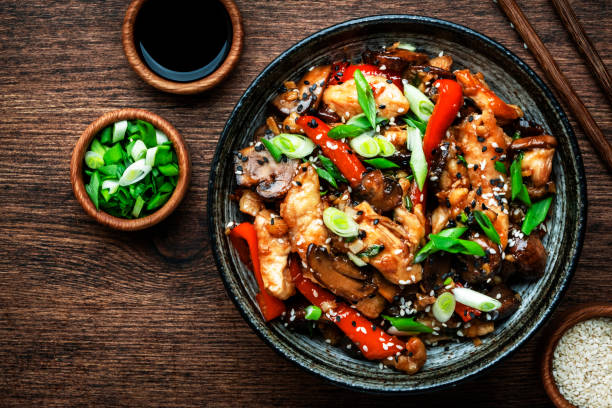Stir fry chicken with paprika, mushrooms, green chives and sesame seeds in ceramic bowl.  Asian cuisine dish. Wooden kitchen table background, top view stock photo