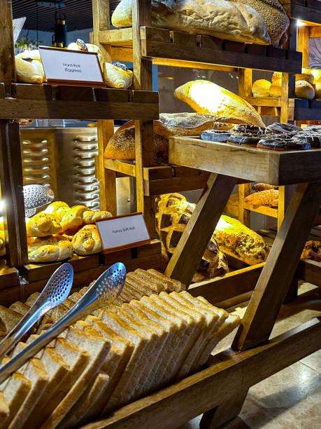 istock Image of hotel breakfast buffet display on wooden shelving unit, rows of bread slices, white loaves, danish pastries, glazed ring doughnuts and cakes, metal serving tongs, focus on foreground 1479934101