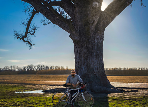 Portrait of Indian American male bicyclist standing by a large oak tree in Midwest in early spring