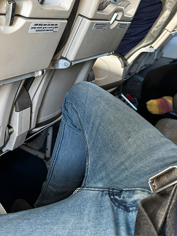 Stock photo showing the interior view of an airplane with a man sat with legs spreads in order to fit into seat.