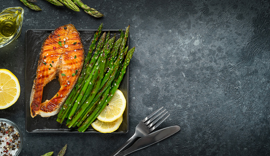 Grilled trout steak and asparagus on grill pan on dark background. Copy space, top view.