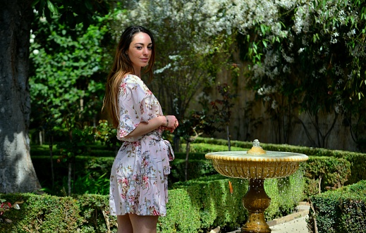 Young brunette woman standing in pink dress in a mediterranean garden near a fountain, Spain, Europe