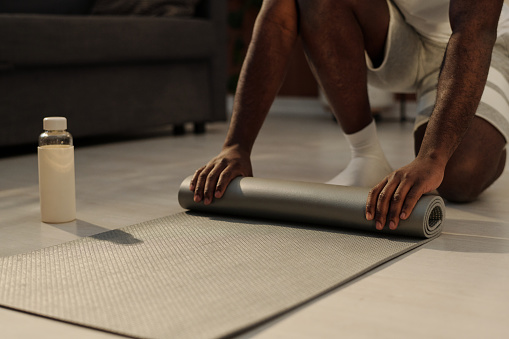 Close-up of hands of young man unrolling grey mat on the floor while going to have morning workout in living room on weekend