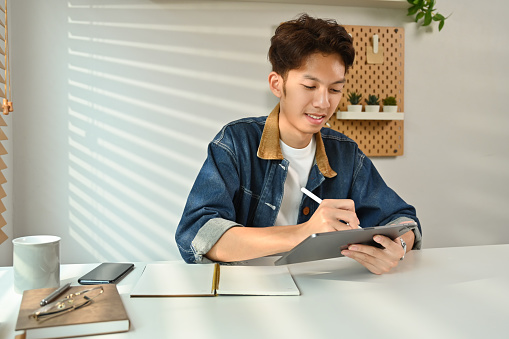 Pleasant asian man freelancer in stylish outfit working online or surfing internet on digital tablet.