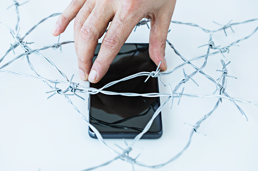 Human hand is trying to touch his smart phone trying not to be injured by barbed wire in front of white background.  Representing news, social media restrictions and  freedom of press and speech.