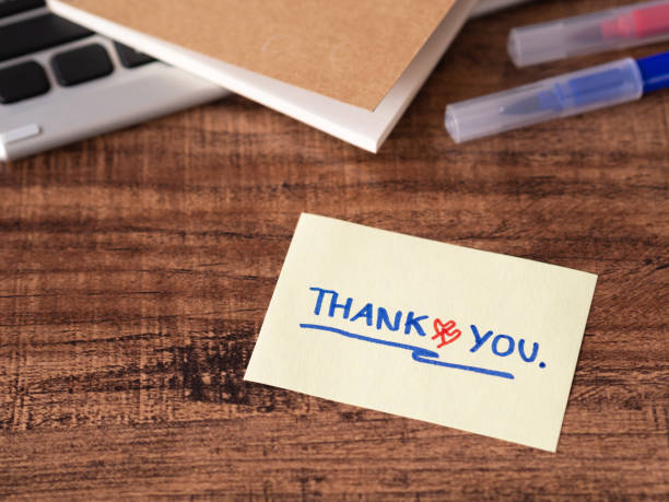 istock Hand writing thank you with red heart on yellow note on wooden table. There is a backdrop as a blur of office equipment. 1479965674