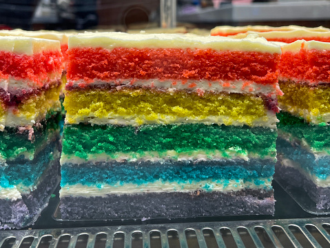 Stock photo showing close-up view of bakery display cabinet of indulgent slices of rainbow cake dessert with multi coloured layers of purple, turquoise blue, green, yellow, and red sponge, decorated with buttercream, available for purchasing by the slice.