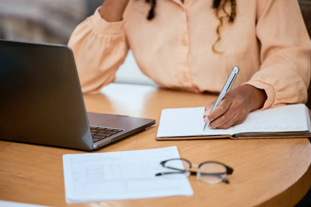 istock Hand, writing and pen with a freelance black woman doing remote work using a laptop in her home. Norebook, documents and computer with a female freelancer doing business in her house or office 1479966408