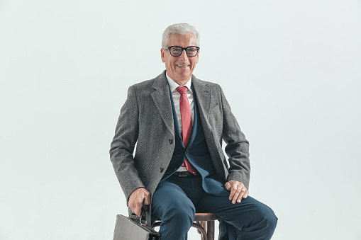 happy ld man with grizzled hair in suit with long coat and suitcase sitting on wooden chair, laughing and posing on grey background