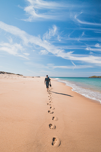 Passionate backpacker, backpack and boots in hand, walks along the Praia da Ilha do Pessegueiro beach on the Atlantic Ocean near Porto Covo, Portugal. In the footsteps of Rota Vicentina.
