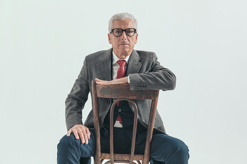 fashion man in his 60s holding arm on chair and posing while sitting on grey background wearing nice suit, with glasses and longcoat