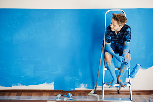 Portrait of teenage boy helping in house renovation. The boy is painting the wall in blue and he is holding a paint roller. 
Shot with Canon R5