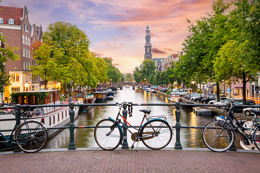 Sunset in Amsterdam - Bicycles parked on a bridge over a canal in the city centre, with typical Dutch houses on the background. Streets illuminated with lights.