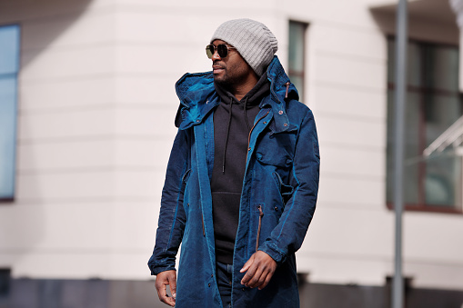 Authentic Portrait of handsome African American man walking in city, wearing stylish outfit parka coat, hoodie, knitted hat and sunglasses. street style fashion male model