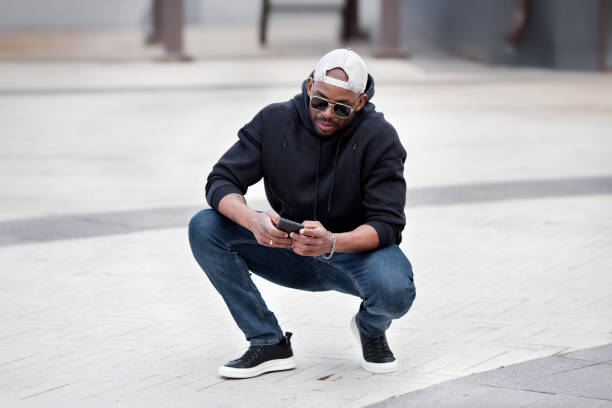 Cool African American man using smartphone, squatting on city street, wears black hoodie, jeans and sneakers, baseball cap and sunglasses. Stylish guy looks at his phone