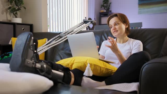 An angry woman with a broken leg is works on a laptop and talking on line. Wide footage