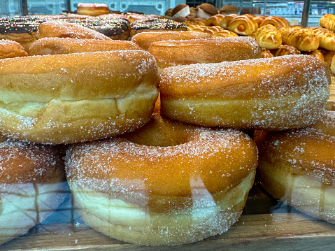 Stock photo showing close-up view of wooden shelf behind a glass panelled bakery display containing a heap of ring doughnuts covered in caster sugar available for purchasing.