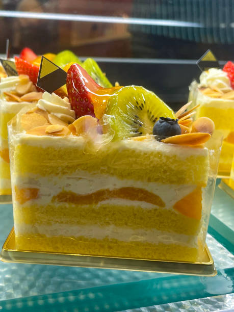 Close-up image of individual lemon cake slice encased in clear plastic triangular box, bakery display of lemon sponge layers separated by buttercream, topped with fruit, kiwi, strawberry and blueberry, focus on foreground