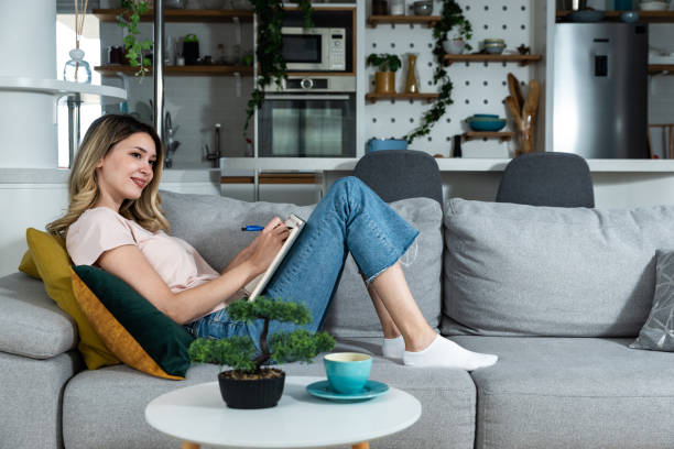 istock Young freelancer woman wedding planner and counselor sitting on sofa drinking coffee and coming up with new ideas for clients as she reads and write what she has written from the wedding plans options 1479996605