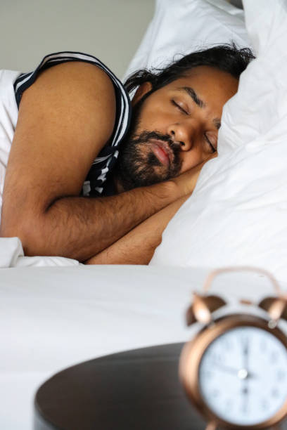 istock Image of bronze metal case retro alarm clock, double bell, analogue display clock with white dial sitting on night stand, Indian man in double bed lying on pillow sleeping on side, focus on foreground 1479999650