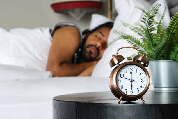 Stock photo showing close-up view of a bronze metal case retro alarm clock with double bell on night stand. An Indian man can be seen lying in the double bed, next to the beside table, sleeping.