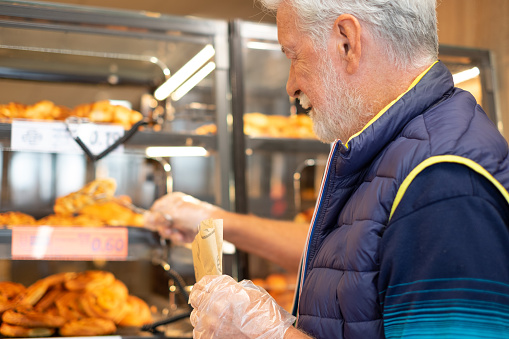 Smiling senior man buying freshly baked pastry in supermarket shopping for groceries paying attention to cost and quality - concept of consumerism, price increase, inflation