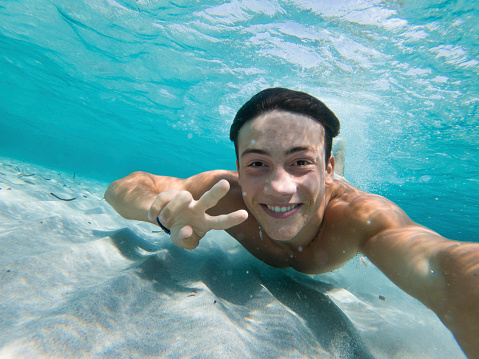 Young boy enjoy summer holiday vacation taking selfie underwater swimming at the sea with sand ground and blue water. Happy tourist swim in crystal clean ocean water doing victory gesture with hands