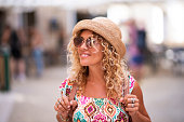 Happy tourist woman with blonde long curly hair and hat looking around and walking alone with backpack enjoying outdoor leisure activity and holiday vacation time. People defocused in background.