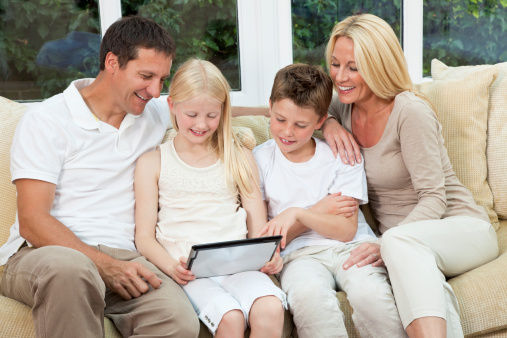 An attractive happy, family of mother, father, son and daughter sitting on a sofa at home having fun using a tablet computer