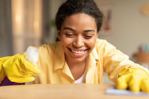 Happy black woman with sprayer detergent and rag cleaning table, lady wearing rubber gloves tidying home, closeup. Doing domestic chores