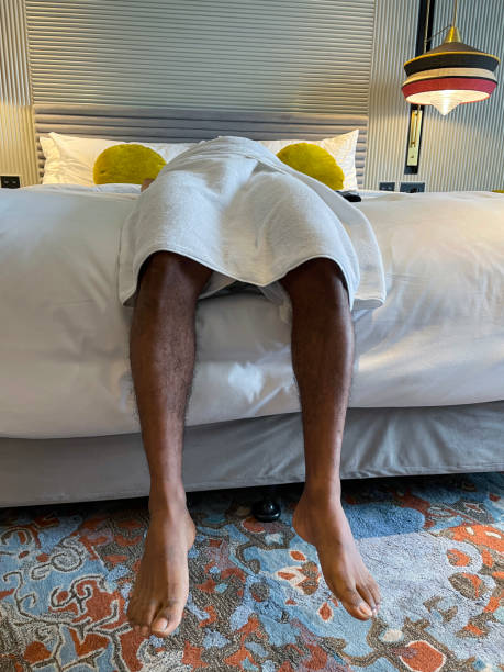 ホテルの部屋の真ん中に丸い腰を巻いた白いタオルを持つ認識できない男性の接写画像が、ダブルベッドの羽毛布団の寝具、ベッドからぶら下がっている脚、前景に焦点を合わせている - double bed headboard hotel room design ストックフォトと画像